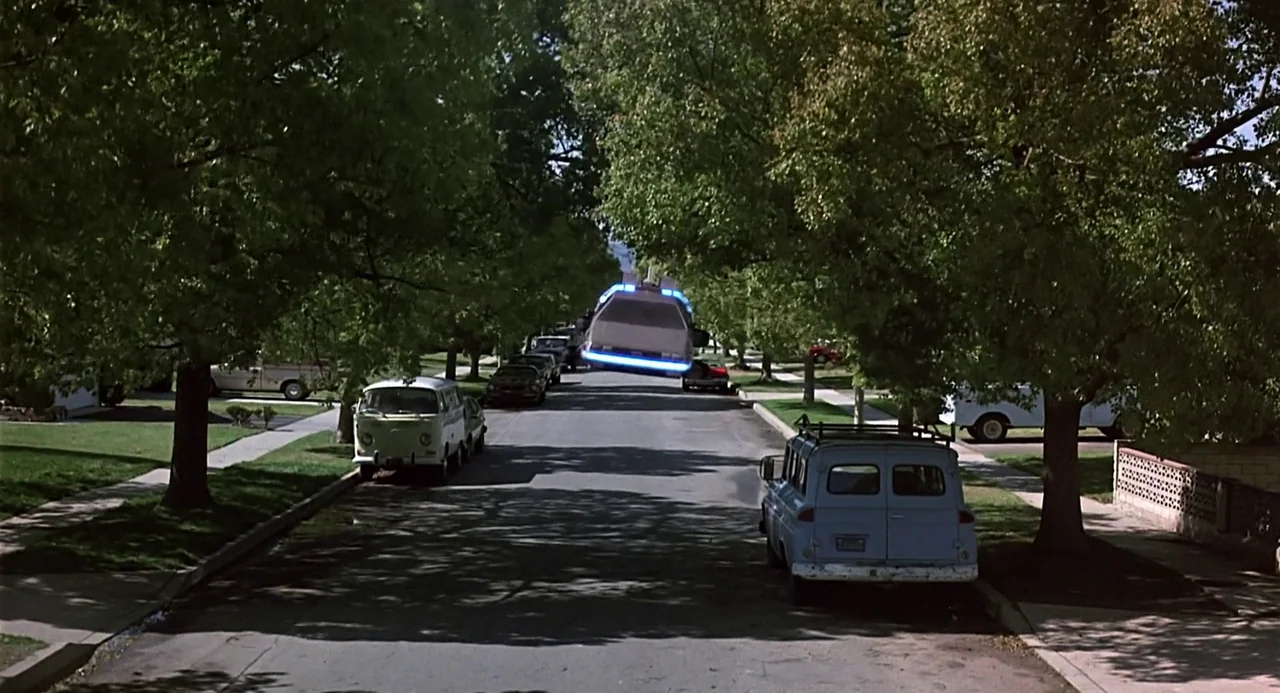Back to the Future (1985) movie still: wide — A quiet suburban street with mature trees lining the sidewalks and parked cars. A silver DeLorean car…