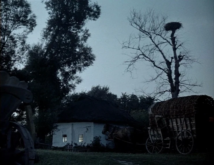 Viy (1967) movie still: wide — A rural setting features a small white farmhouse with glowing windows at twilight. A horse-drawn wagon sits in the…