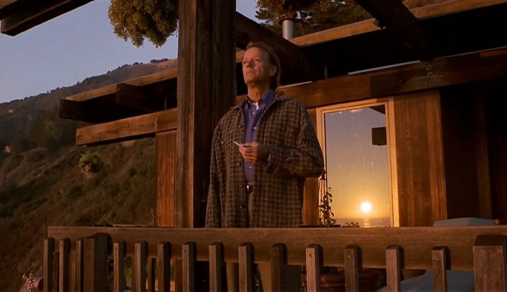 The Limey (1999) movie still: medium — A wooden balcony attached to a house overlooks a coastal hillside at sunset. A man stands on the balcony,…