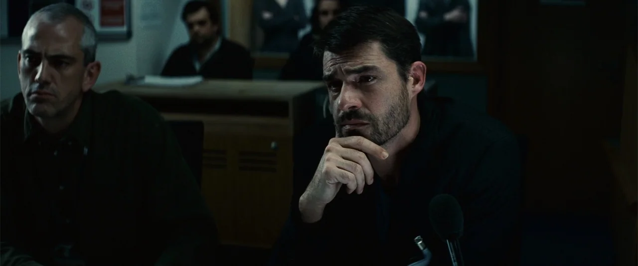 Hereafter (2010) movie still: medium — A dimly lit room with a desk and microphone setup indicates a formal environment. Two men sit side by side…