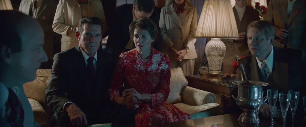 Reagan (2024) movie still: medium — A dimly lit hotel room contains several people dressed in formal 1970s attire. A middle-aged couple sits on a…
