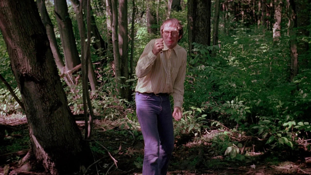 I Spit On Your Grave (1978) movie still: medium — A dense patch of woods with thin, tall trees and thick green undergrowth. A man stands in the…