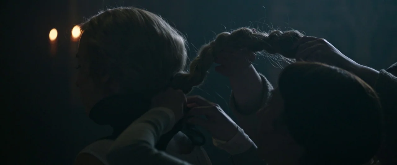 The Last Duel (2021) movie still: closeup — A dimly lit room with stone walls shows two women in a close-up. One woman braids the blonde hair of…