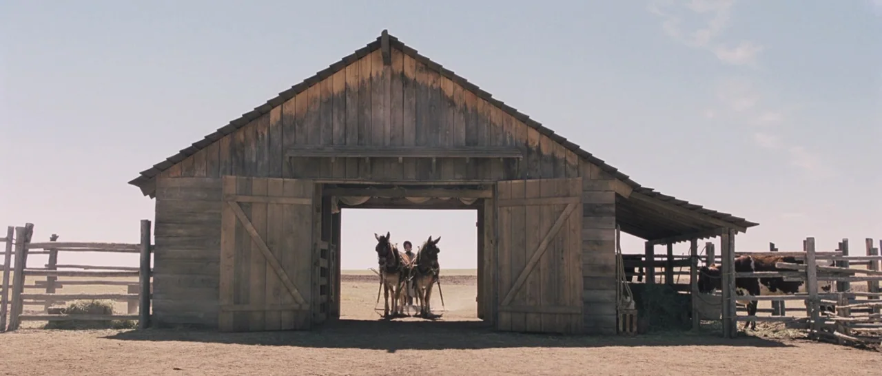The Homesman (2014) movie still: wide — A weathered wooden barn sits in the middle of a sparse, dry open field. A child stands between two mules…