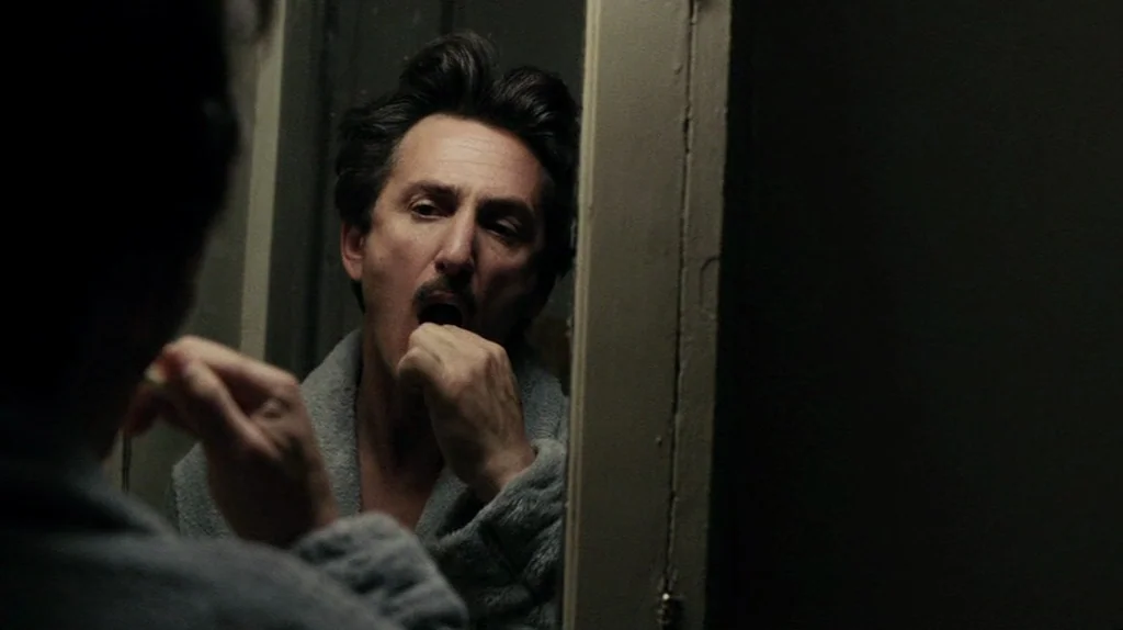 The Assassination of Richard Nixon (2004) movie still: closeup — A dimly lit bathroom setting features a man standing before a mirror. The man…