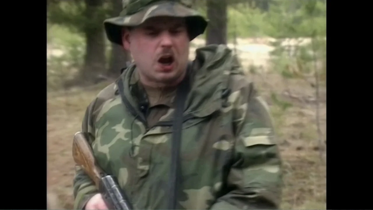 The Forever Prisoner (2021) movie still: medium — A man in a camouflage jacket walks through a sparsely wooded area outdoors. He is shouting with…