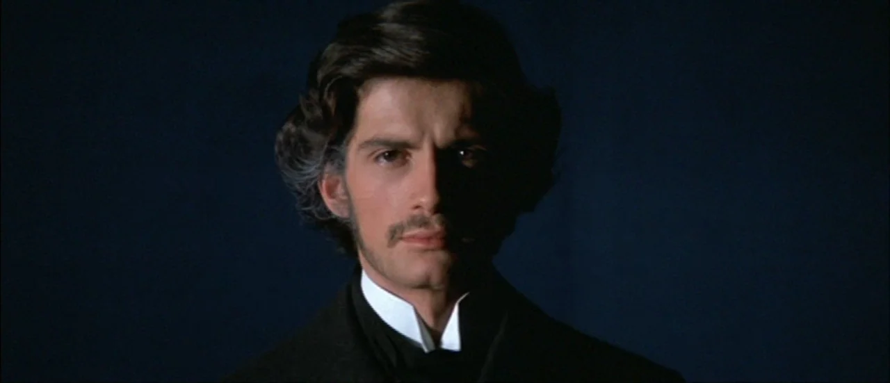 Ludwig (1973) movie still: closeup — A man stands against a dark, featureless backdrop with minimal lighting. He faces forward while his face is…