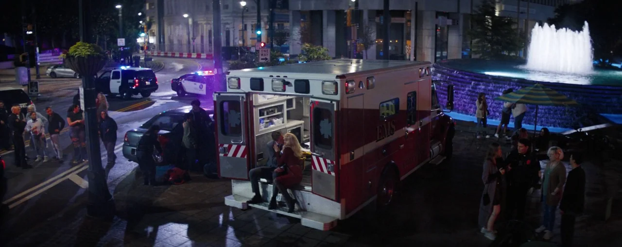 The Naked Gun (2025) movie still: wide — A nighttime city plaza features a lit water fountain and an ambulance parked nearby. A man and a woman sit…