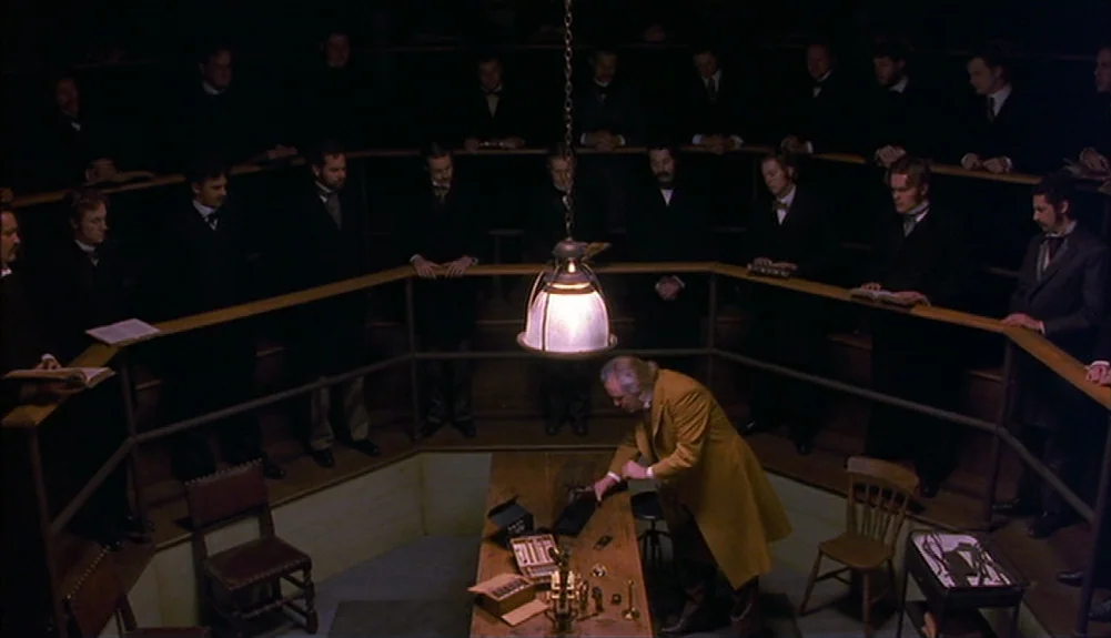 Bram Stoker’s Dracula (1992) movie still: wide — A dark wooden lecture hall with tiered semicircular seating is filled with men in suits. A single…