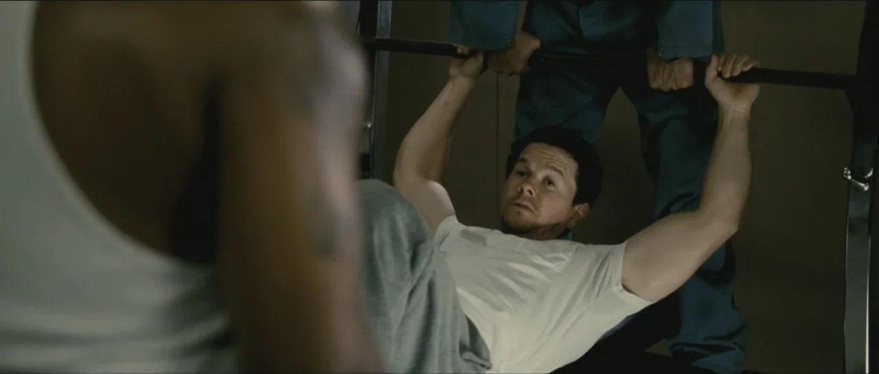 Contraband (2012) movie still: medium — A sparse room with metallic weightlifting equipment features two men engaged in a workout. One man lies on a…