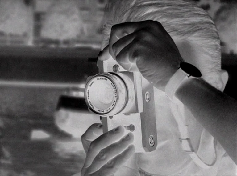 Une Femme Mariée (1964) movie still: closeup — A man holds a vintage Nikon camera to his eye at an outdoor waterfront location. The image is…