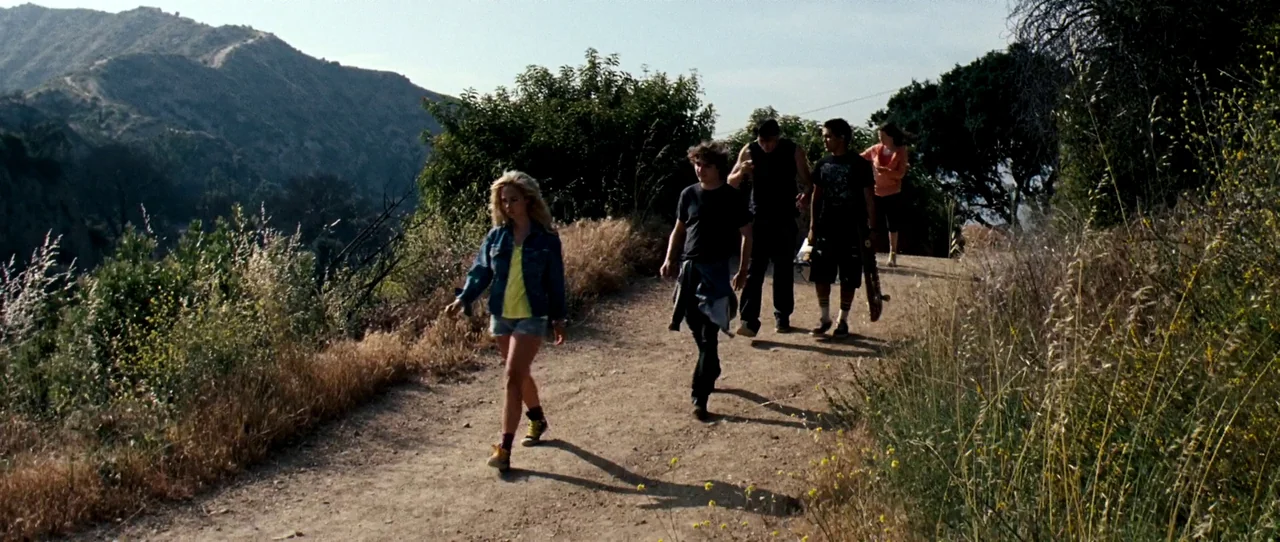 Little Birds (2011) movie still: fullbody — A group of teenagers walks along a dry dirt path in a hilly landscape. The trail is lined with tall dry…