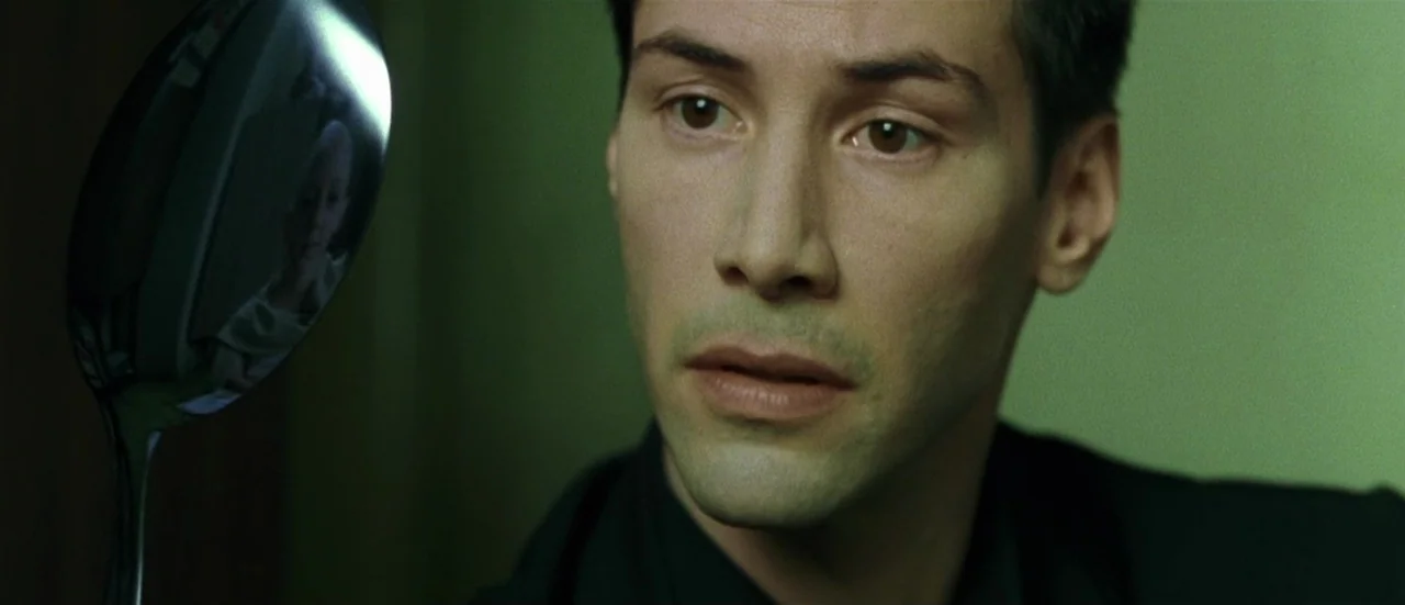 The Matrix (1999) movie still: closeup — A dimly lit interior space with a persistent green tint. A man gazes intently at the reflection in a curved…