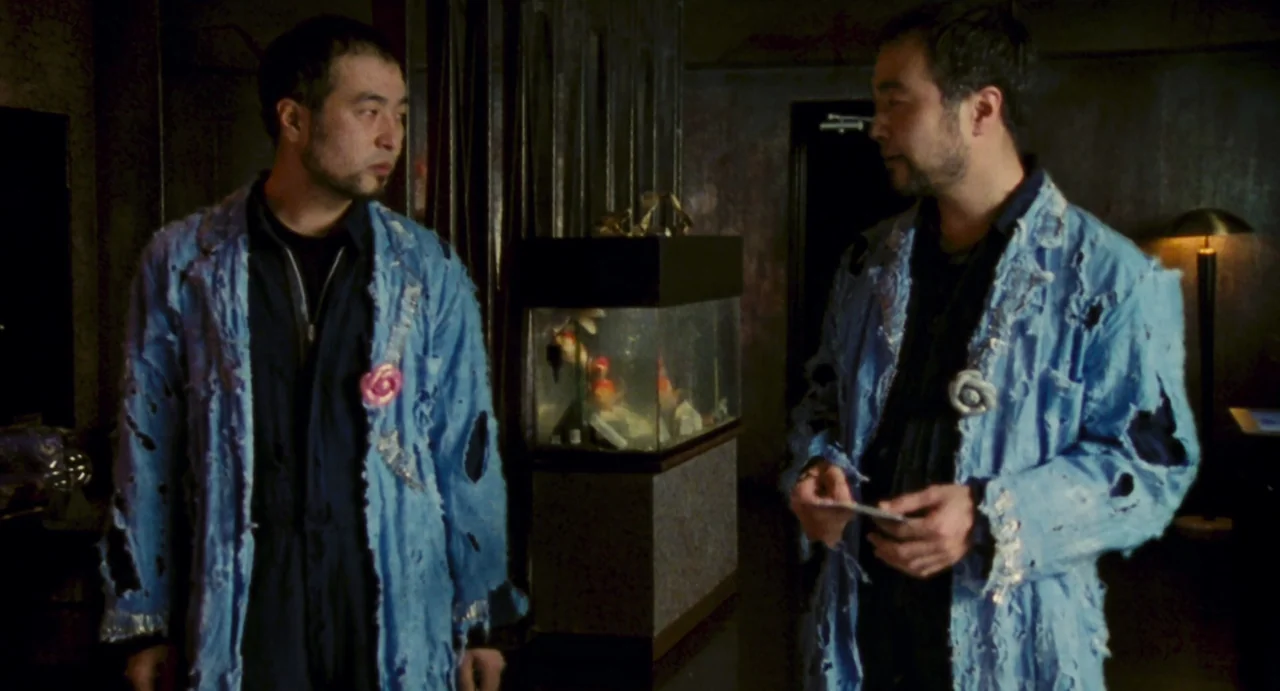 Ichi The Killer (2001) movie still: medium — A dimly lit interior space contains a rectangular fish tank between two identical men. Both men wear…
