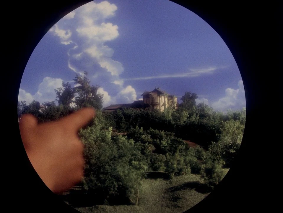 House (Hausu) (1977) movie still: wide — A rural hillside features a small residential building nestled among dense green trees. An out-of-focus…
