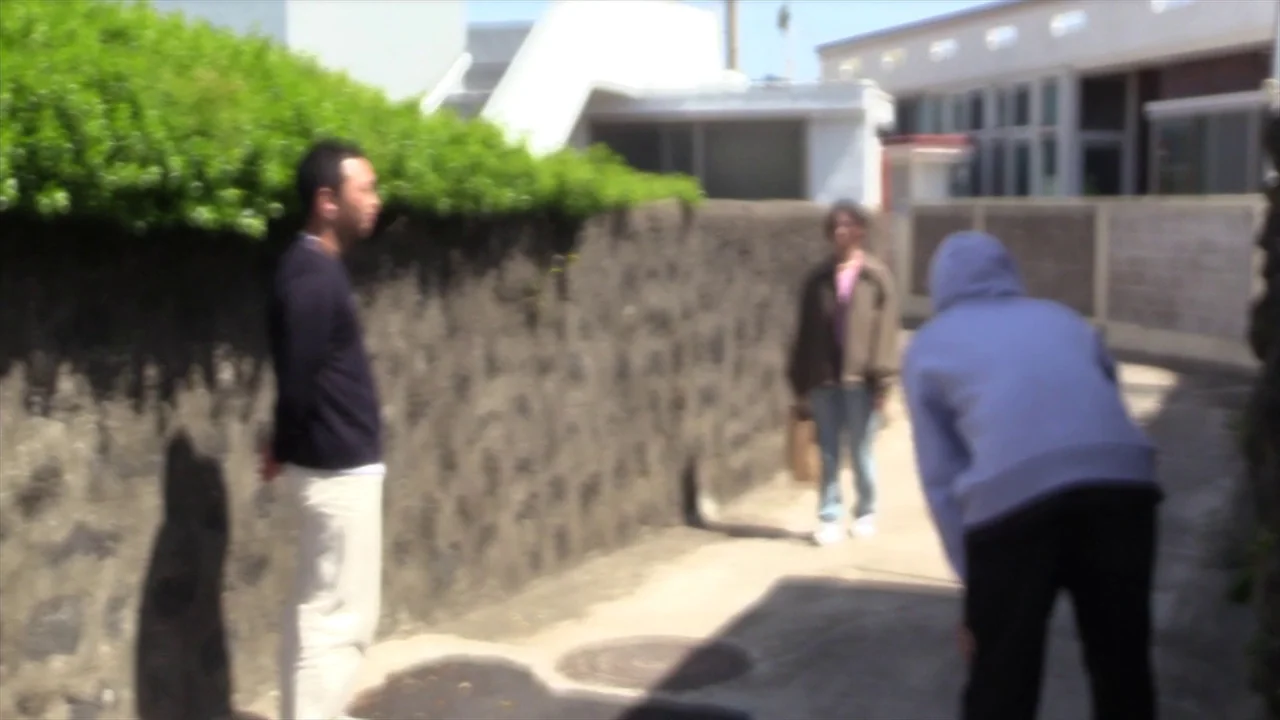 In Water (2023) movie still: fullbody — A narrow outdoor pathway is flanked by a rough stone wall with green hedges. Two men and one woman stand on…