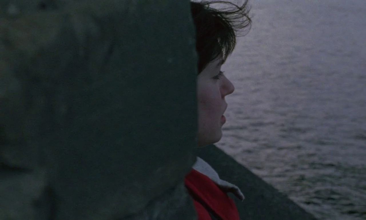 Rosetta (1999) movie still: closeup — A person stands beside a concrete pillar overlooking a dark body of water. The subject is captured in profile…