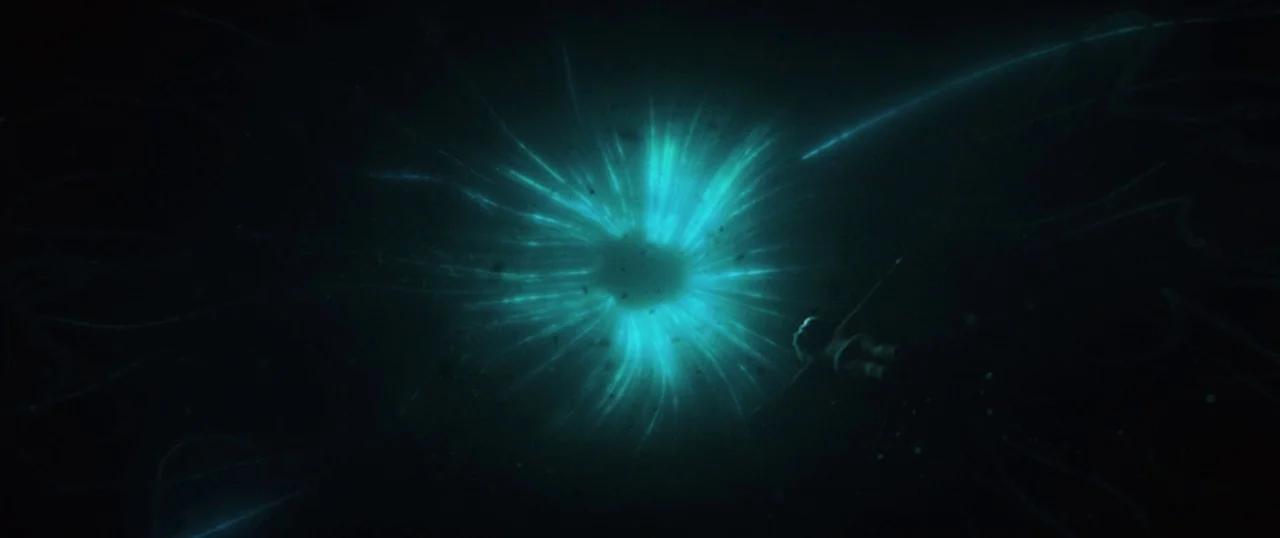 Sea Fever (2019) movie still: wide — A dark, expansive underwater environment is dominated by a central, pulsing blue light. A small, indistinct…