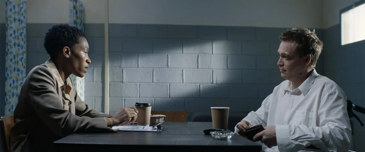 Dogman (2023) movie still: medium — An interior interview room features grey cinder block walls and a simple dark table. A woman and a man sit…