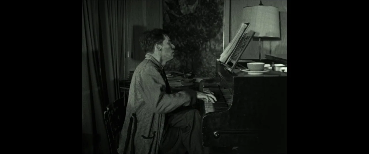The House That Jack Built (2018) movie still: medium — A dimly lit room contains an upright piano and a small lamp. A man sits at the piano playing…