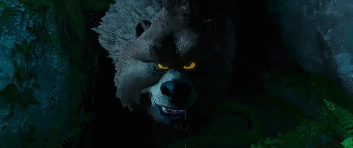 The Wild Robot (2024) movie still: closeup — A large, dark, furry bear with glowing yellow eyes peers out from a cavern entrance. The bear is…