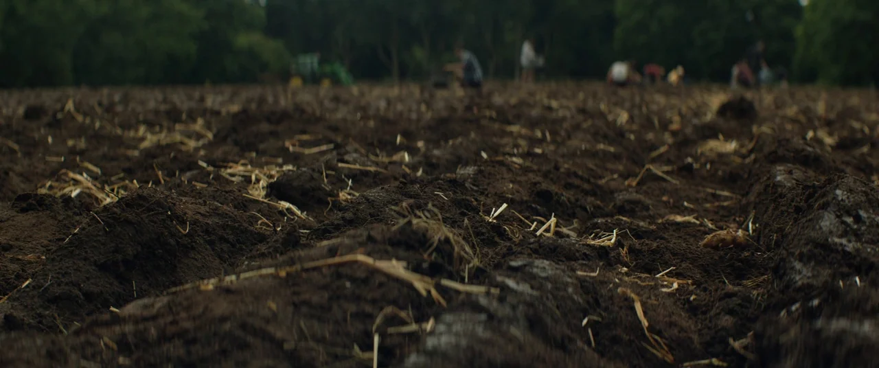 Beast (2017) movie still: wide — A field of freshly tilled dark brown soil with scattered straw and dry stalks. Blurred silhouettes of multiple…