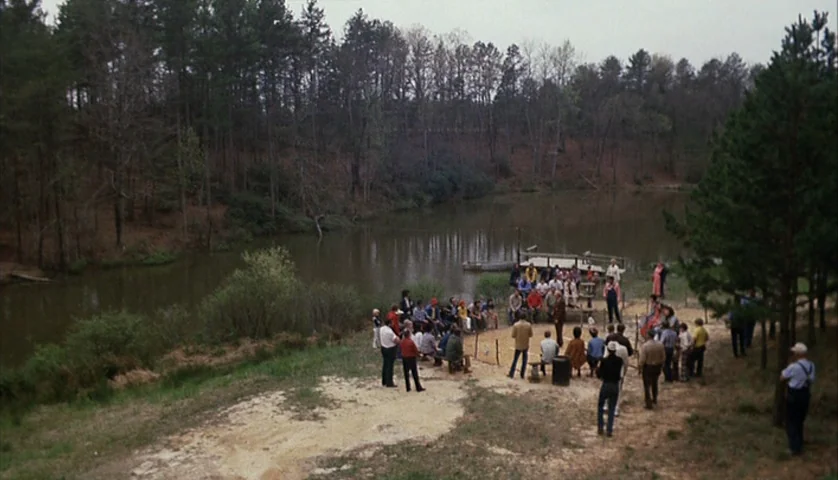 Cockfighter (1974) movie still: wide — A group of people gathers in a wooded area beside a calm, murky lake. The crowd stands and sits in a loose…