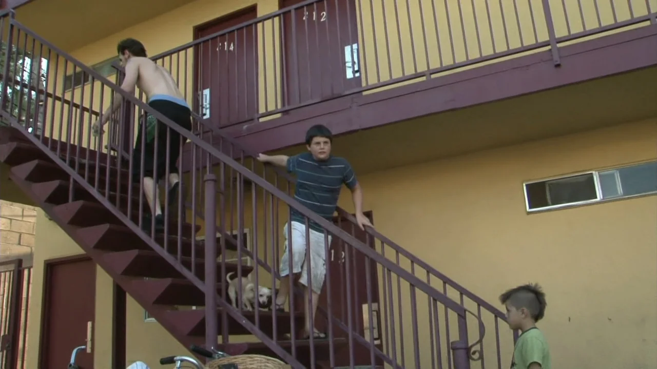 Homeless: The Motel Kids of Orange County (2010) movie still: fullbody — A yellow stucco motel building features a dark red painted metal staircase…