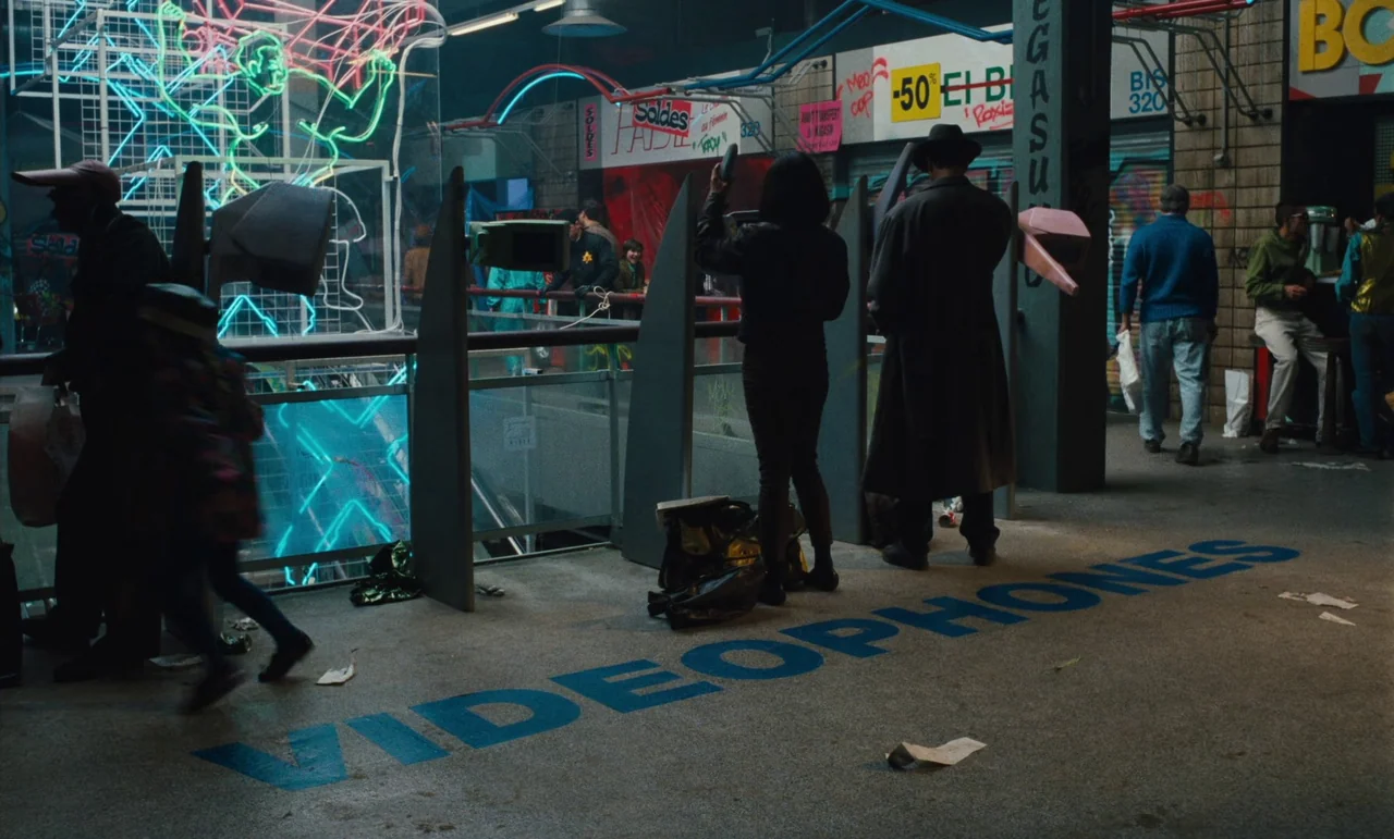Until The End Of The World (1991) movie still: wide — A dimly lit, multi-level concrete shopping mall concourse features blue 'VIDEOPHONES'…