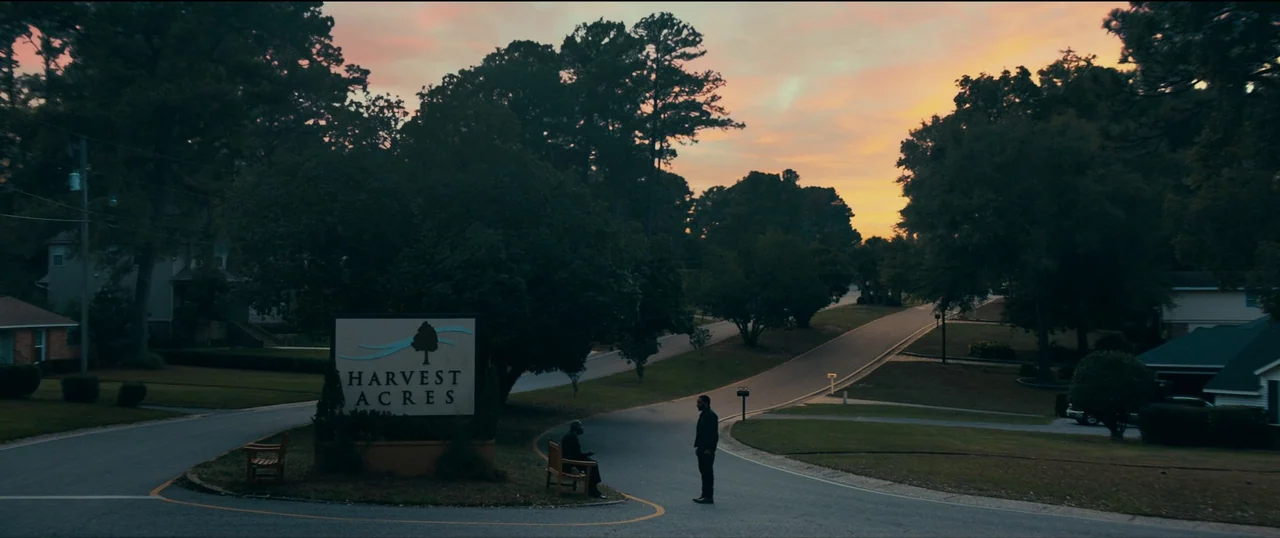 The Life of Chuck (2024) movie still: wide — A suburban entrance sign reading Harvest Acres sits at a paved road intersection. One man stands while…