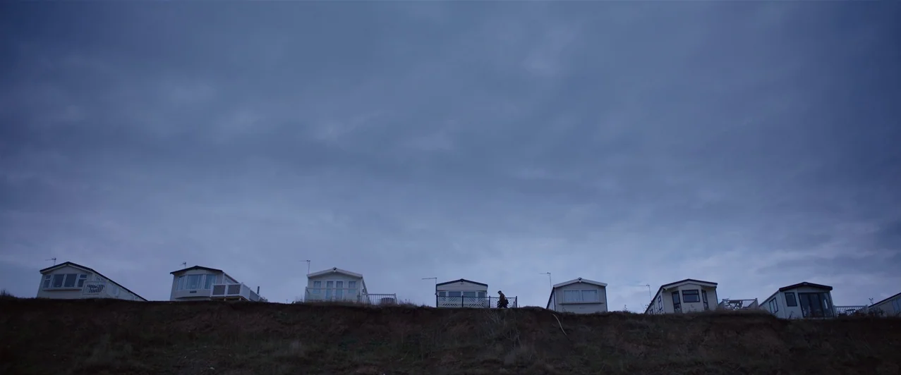 Ghost Stories (2017) movie still: wide — A row of static residential trailers sits along the crest of a grassy cliffside. A lone person walks past…