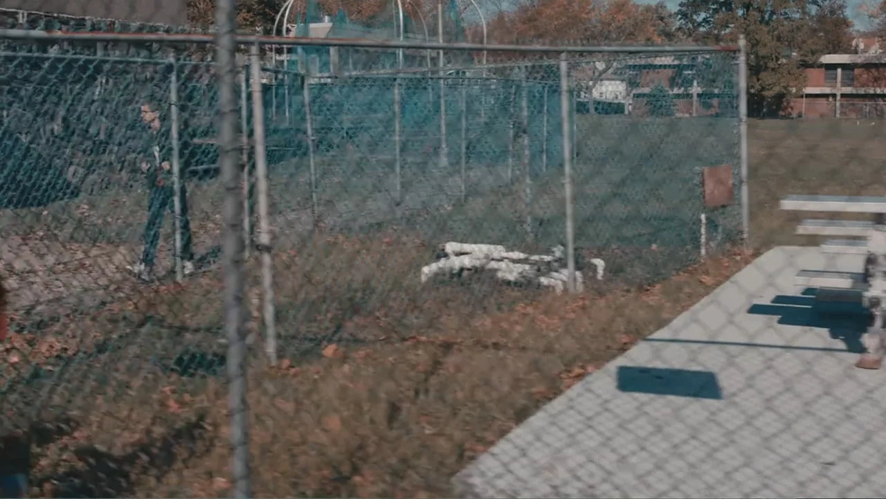Terrifier 2 (2022) movie still: wide — An outdoor park area enclosed by a chain-link fence with a concrete walkway. A person in dark clothing walks…