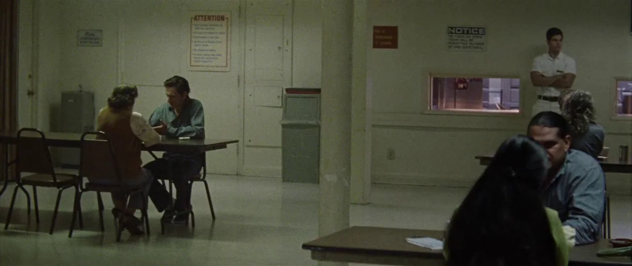 Low Down (2014) movie still: wide — A utilitarian institutional space with stark white walls and multiple tables for dining. A man and woman sit…