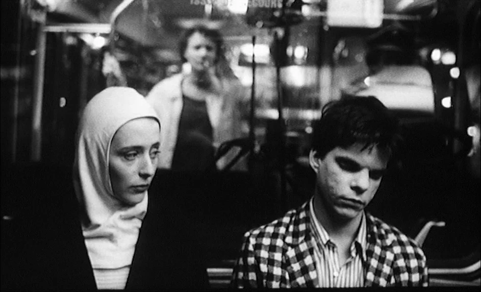 Boy Meets Girl (1984) movie still: medium — A dark, dimly lit bus cabin at night with a blurred passenger visible behind. A woman in a white hood…
