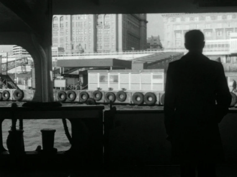 Death and Transfiguration (1983) movie still: medium — A man stands on a ferry deck looking out toward a city waterfront. The background shows…