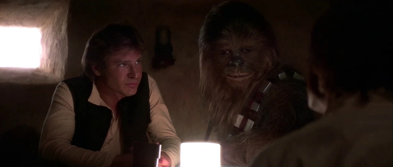 Star Wars (1977) movie still: medium — A dimly lit, textured space features a glowing lamp centered on a small table. A man and a tall, fur-covered…