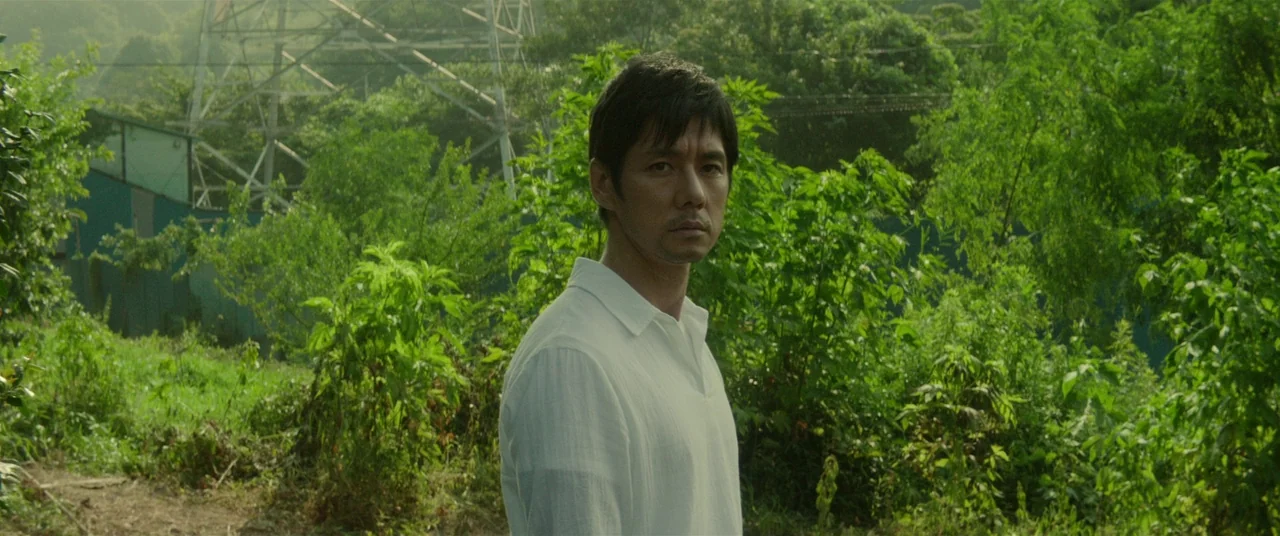 Creepy (2016) movie still: medium — A dense green area features overgrown vegetation and a tall metal electrical tower. A man in a light shirt…