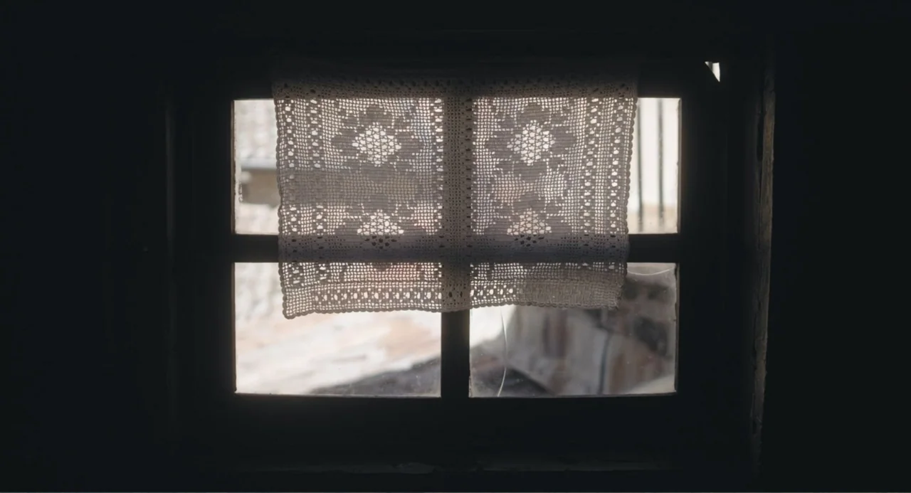 Los días azules (2025) movie still: closeup — A dark room with a small paneled window looking out to a bright exterior. A white lace doily hangs…