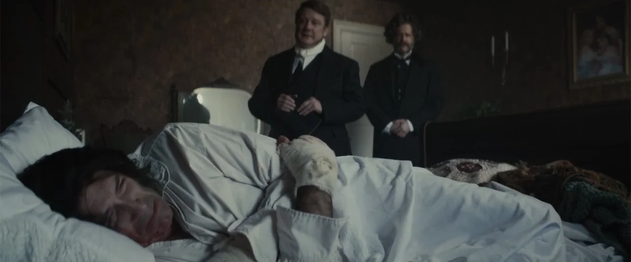 I Heard the Bells (2022) movie still: medium — A dimly lit 19th-century room features ornate wallpaper and a bed with white linens. A man with…