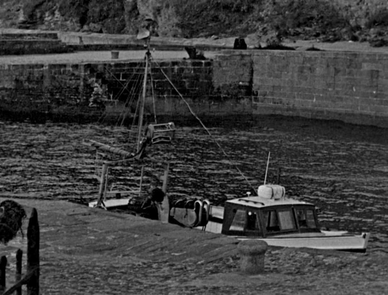 Bait (2019) movie still: wide — A small white fishing boat floats in a quiet harbor beside a stone wall. The water ripples against the concrete pier…