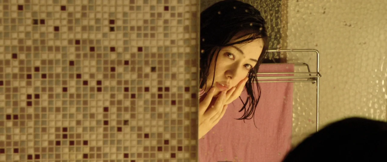 Norwegian Wood (2010) movie still: closeup — A tiled surface sits in the foreground beside a mirror reflecting a young woman. The woman touches her…