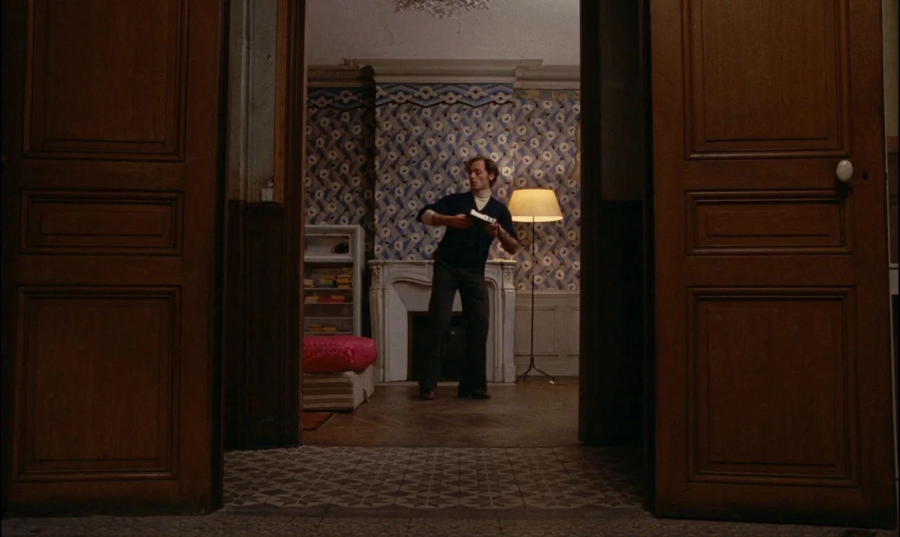 Serie Noire (1979) movie still: fullbody — An interior space features patterned blue wallpaper, a marble fireplace, and tall wooden double doors. A…