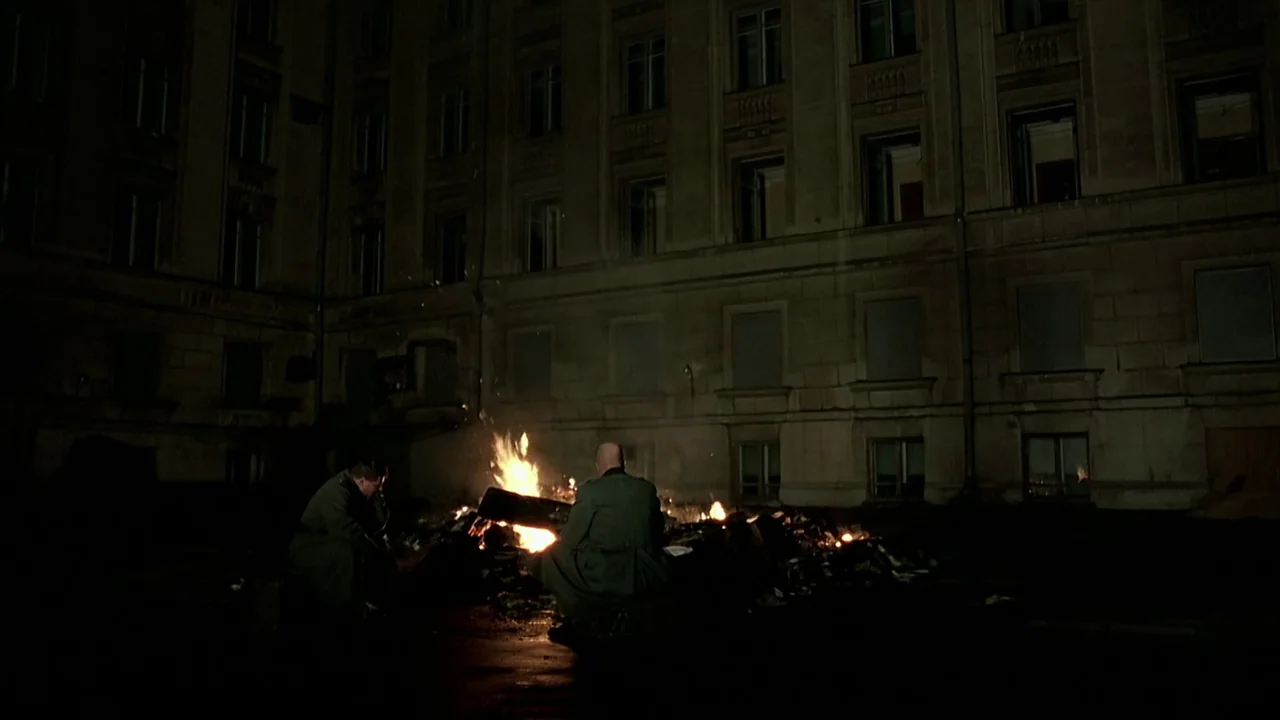 Downfall (2004) movie still: wide — A dark, expansive urban courtyard is surrounded by a large, multi-story masonry building. Two men sit on the…