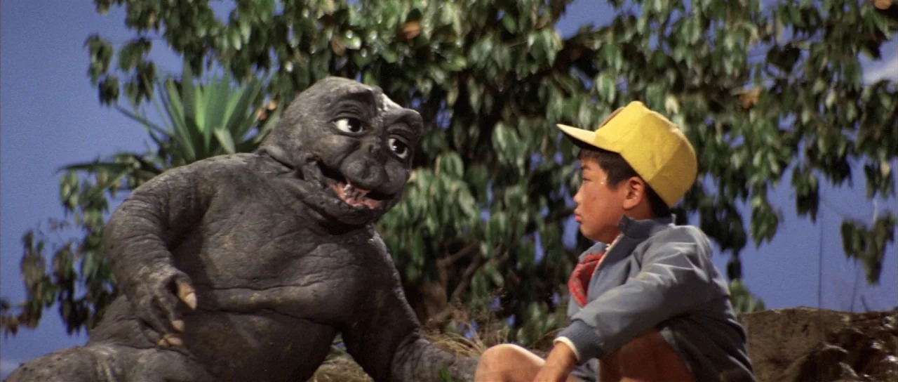All Monsters Attack (1969) movie still: medium — A dark, rubbery creature sits beside a young boy on a rocky hillside. Lush green trees stand in the…