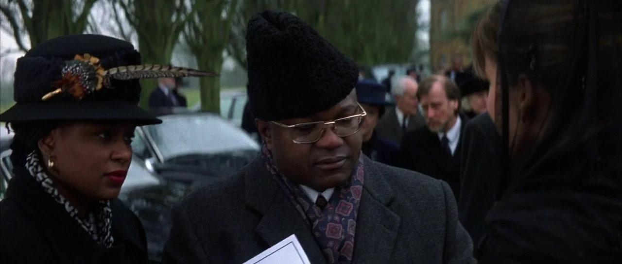 The World is Not Enough (1999) movie still: medium — An outdoor gathering with several people standing near parked cars under tall bare trees. A man…