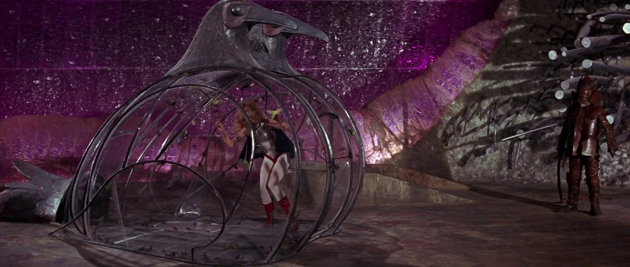 Barbarella (1968) movie still: wide — A surreal chamber with metallic walls illuminated by vibrant purple light and dark shadows. A woman is trapped…