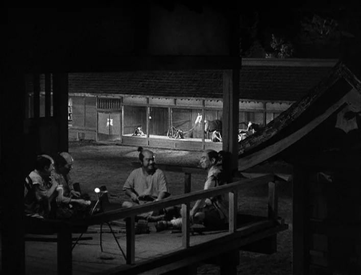 Throne of Blood (1957) movie still: wide — A wooden veranda at night overlooks a stable area with horses in stalls. Four men sit on the porch floor…