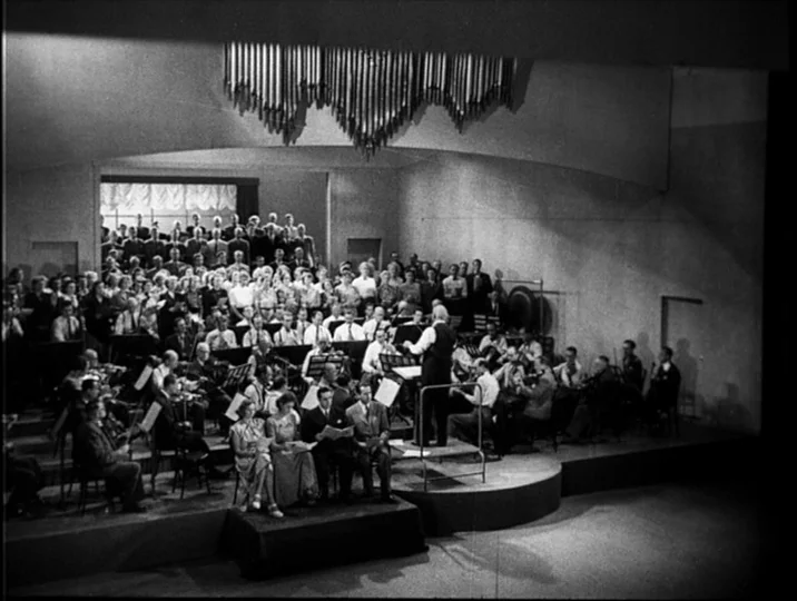 To Joy (1950) movie still: wide — A spacious auditorium features a tiered platform hosting a large orchestra and choir. The conductor stands at the…