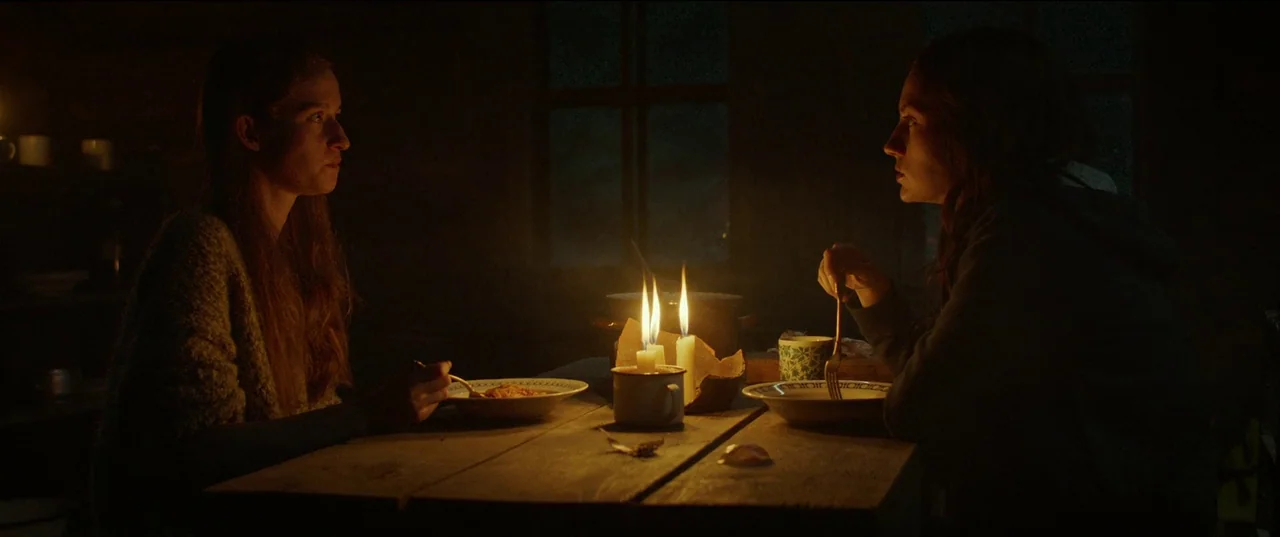 Nightsiren (2022) movie still: medium — A rustic room is illuminated solely by three candles centered on a wooden table. Two women face each other…