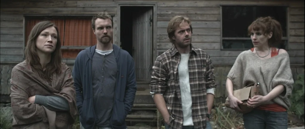 One Hundred Mornings (2009) movie still: fullbody — Four people stand in a horizontal line in front of a rustic wooden building. The figures occupy…